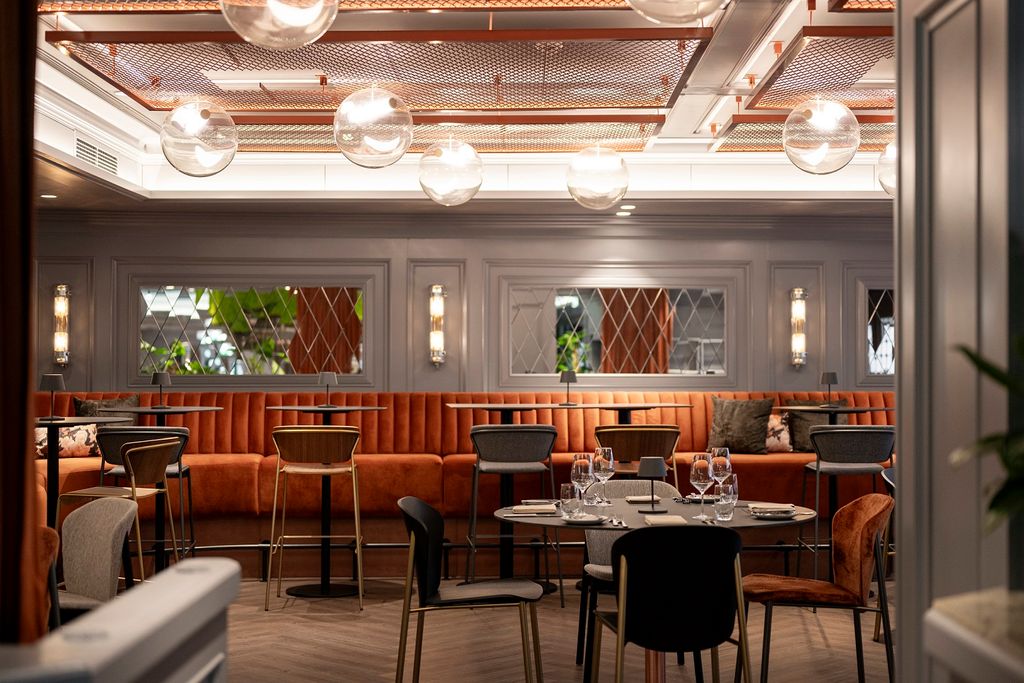 A cozy restaurant interior with modern decor, featuring round tables, cushioned chairs, a long booth with orange upholstery, and spherical ceiling lights.